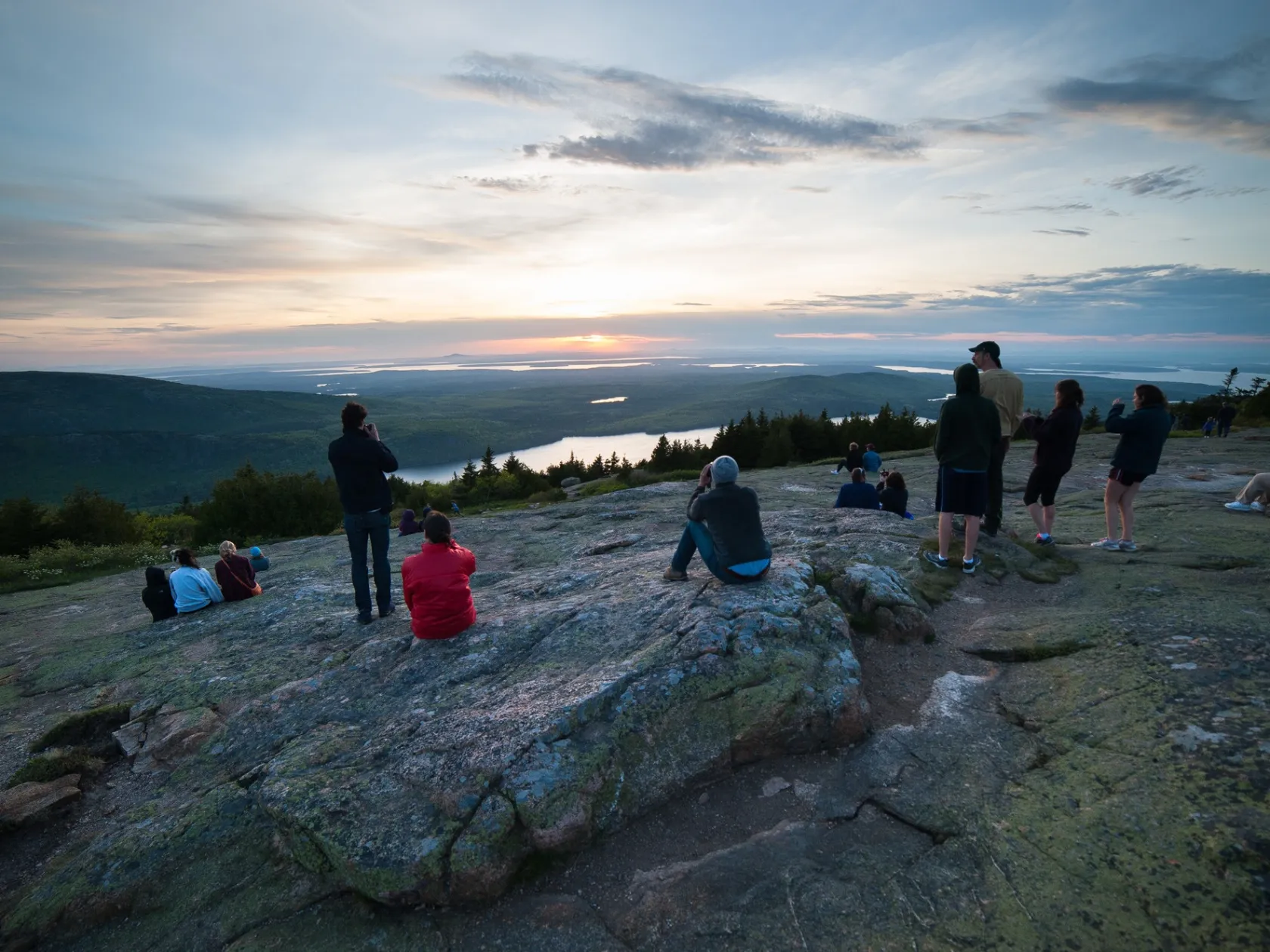 People sitting on a rocky hilltop watching a sunset over distant hills and water.