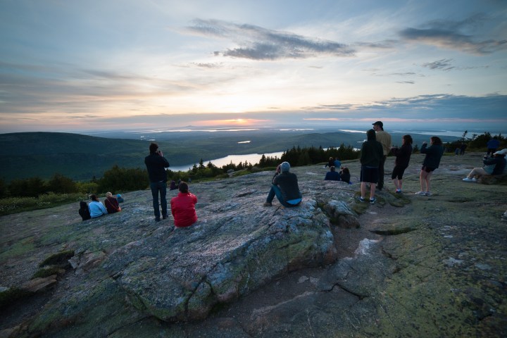 People sitting on a rocky hilltop watching a sunset over distant hills and water.
