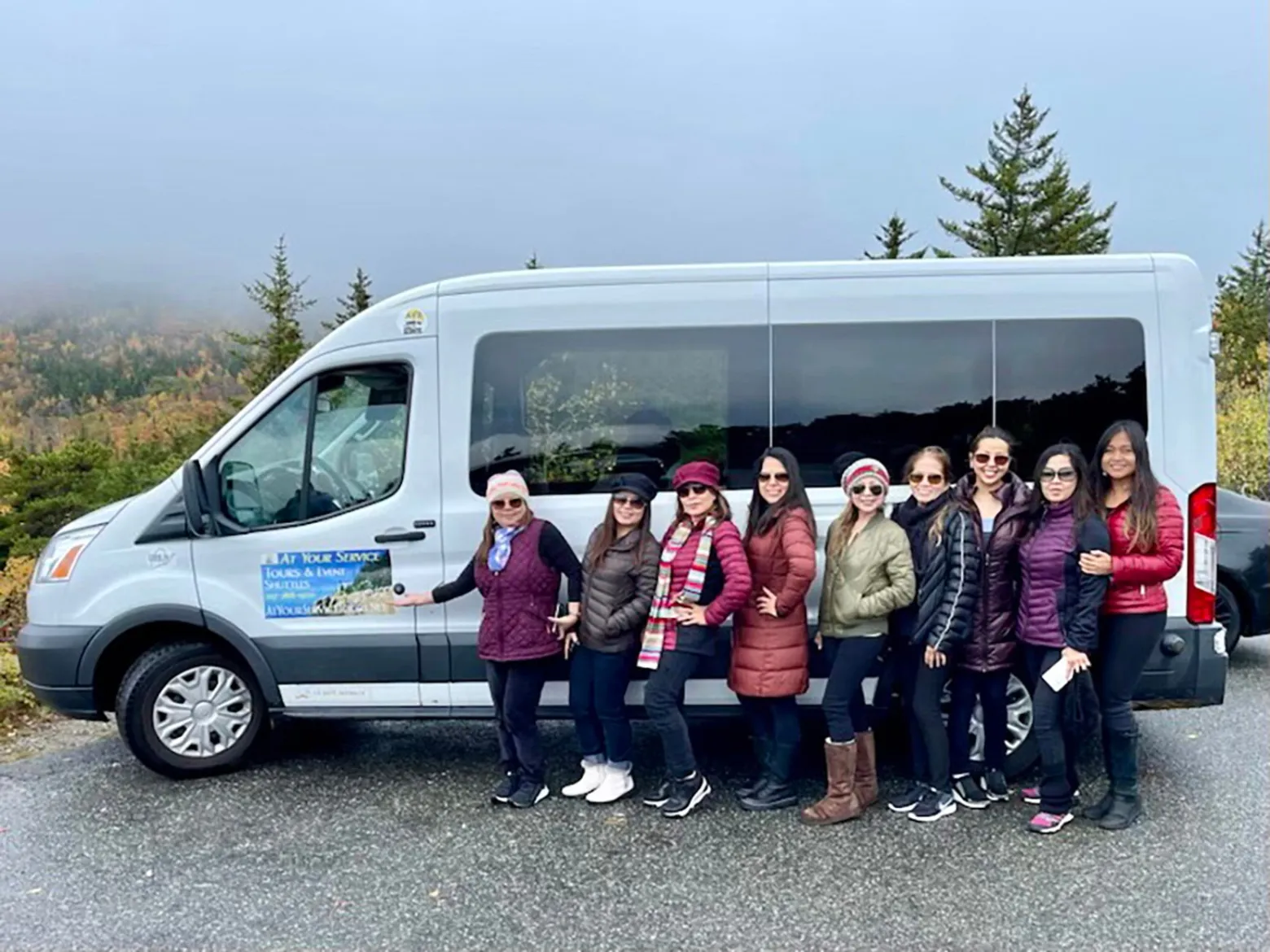 Group of people posing beside a gray van with trees and foggy background.