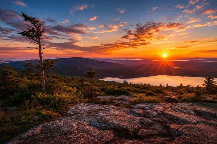 Sunrise over rocky landscape with distant hills, trees, and a glowing sky.