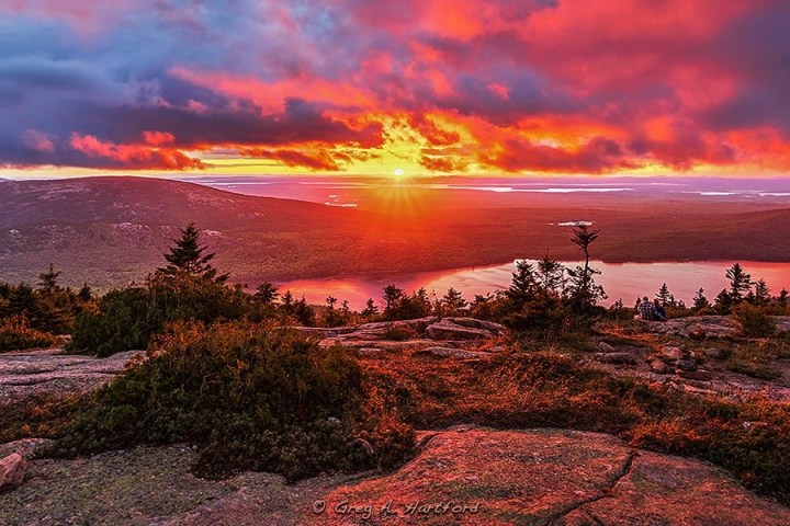 Vibrant sunset over a mountainous landscape with trees and a lake.